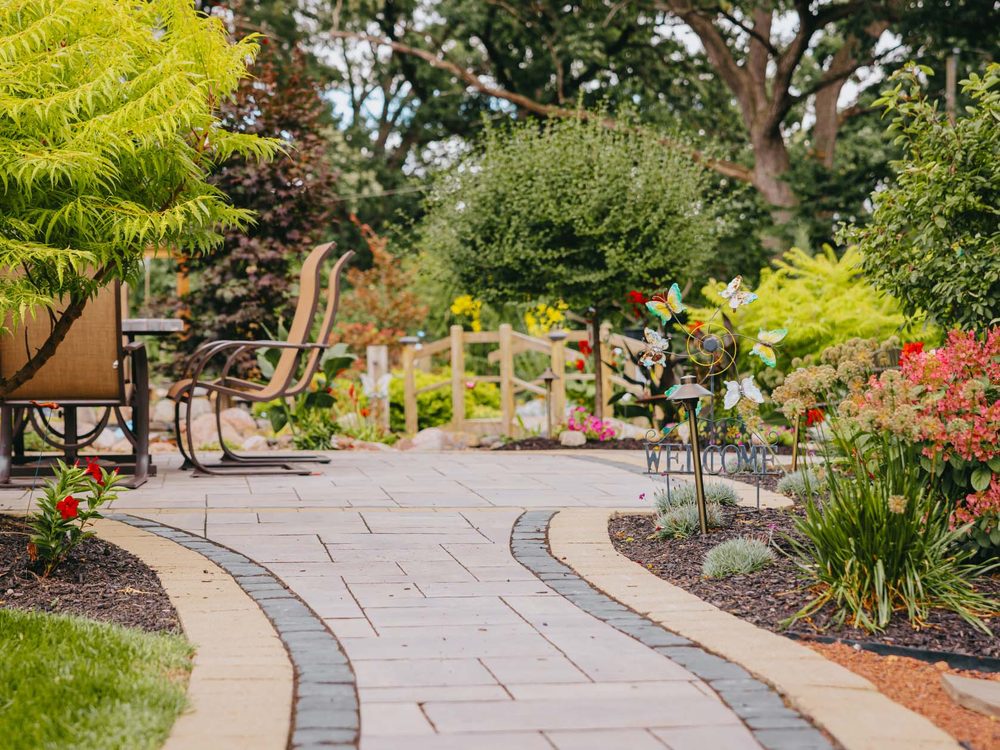 Brick paver walkway curving past a seating area framed by deep green planting.
