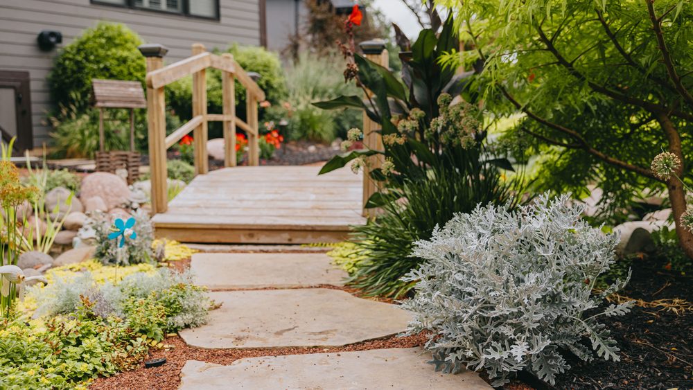 Flagstone walkway leading to a small wooden garden bridge, framed by layered perennial beds.