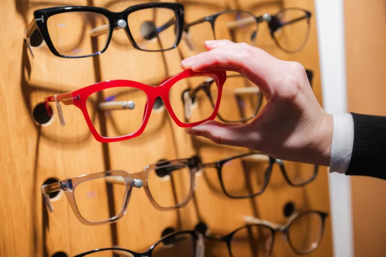 A hand selects red eyeglass frames from a wooden display rack with various glasses.