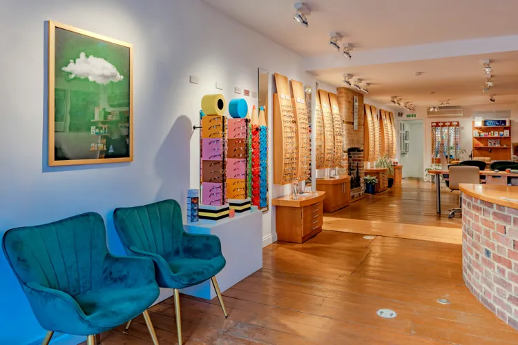 Interior of optician shop with wooden floor, blue chairs, eyeglass displays, and framed cloud artwork.