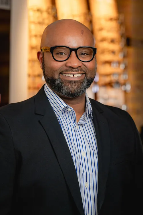 Smiling bald man with glasses, beard, striped shirt, and black blazer against blurred background.