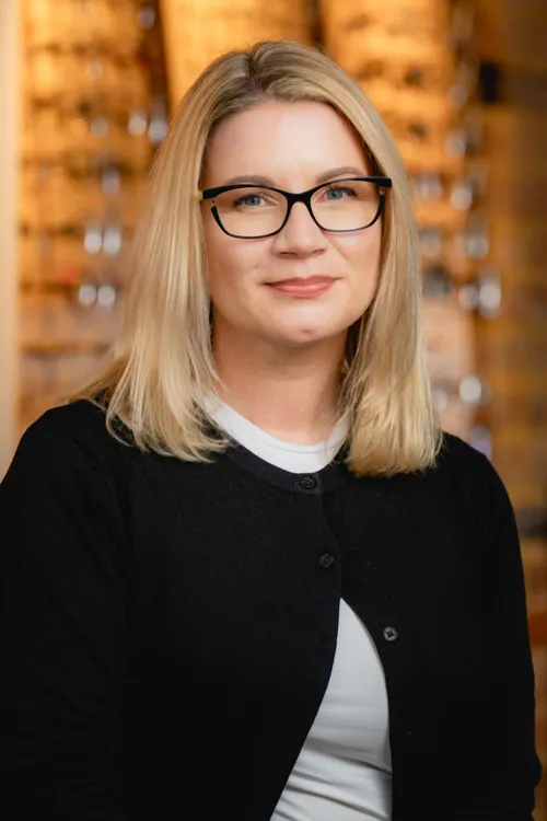 Blonde woman with glasses wearing a black cardigan and a white top, smiling softly with blurred background.