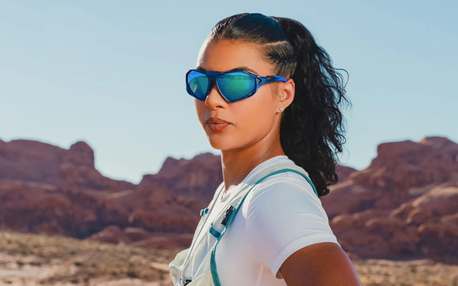 Woman in white shirt wearing reflective blue sports sunglasses in a rocky desert landscape.