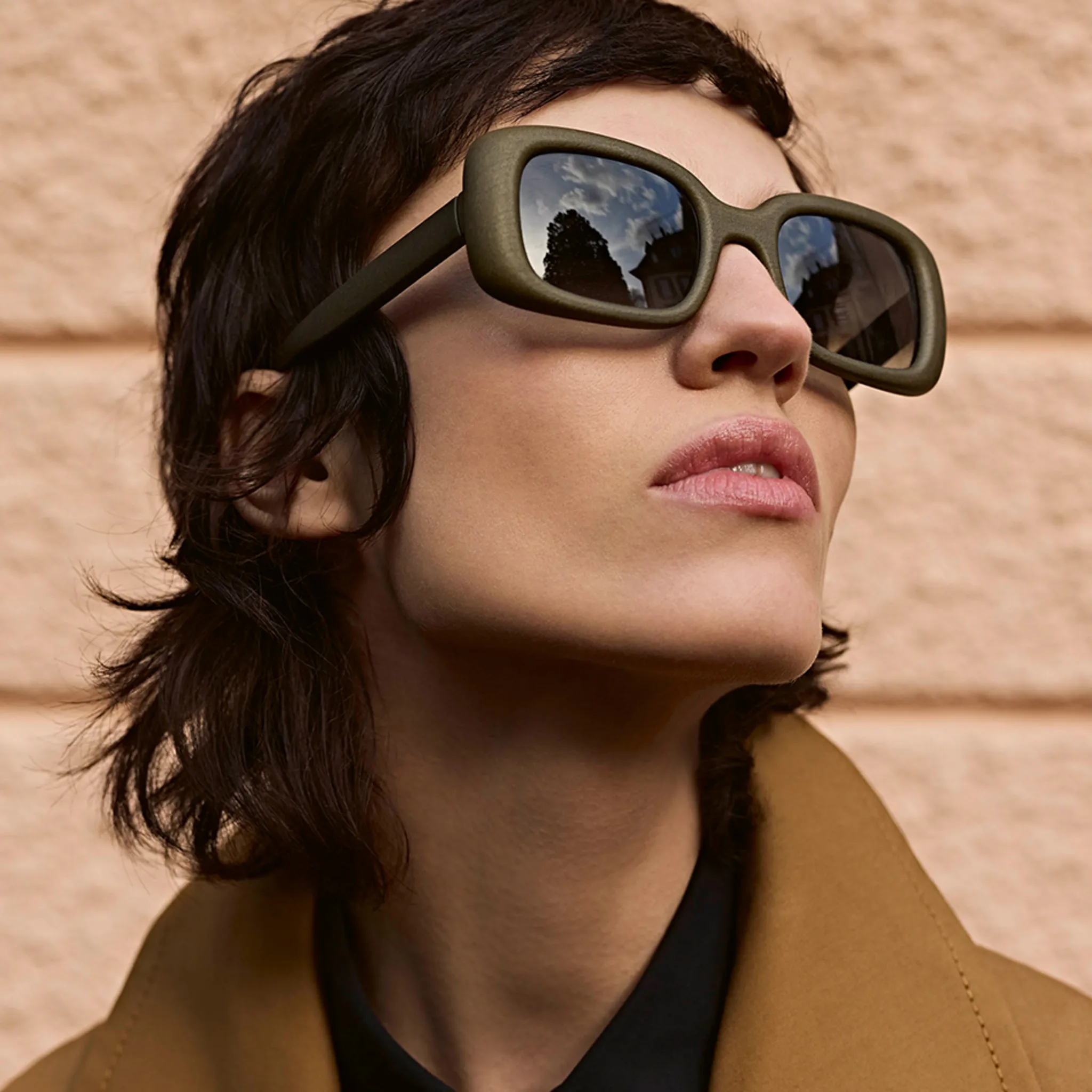 Person with short dark hair wearing large square sunglasses reflecting trees and buildings.
