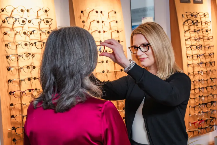 Optician wearing glasses fitting glasses on a woman with gray hair in eyewear shop.