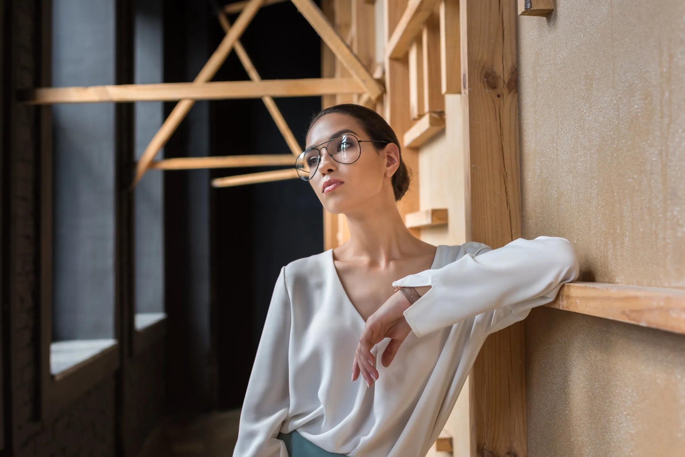Woman with glasses leaning against a wall with wooden geometric decorations in a dim room.