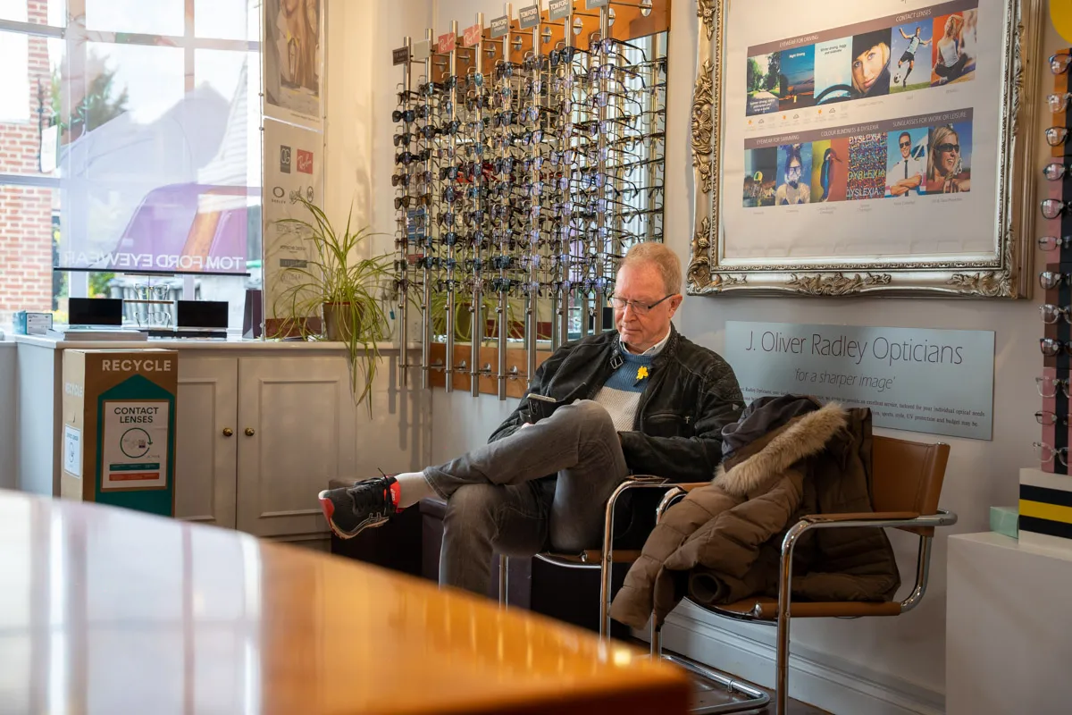 Older man wearing glasses sitting in an optician shop, looking at his phone with eyeglass displays behind him.