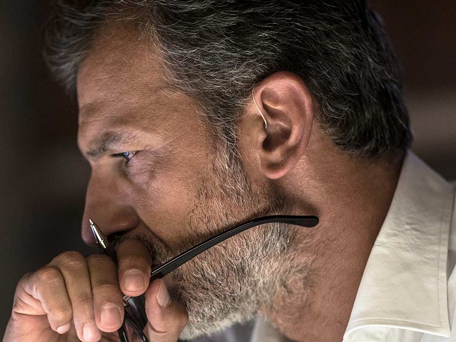 Close-up of a bearded man with a hearing aid deep in thought holding glasses and pen.