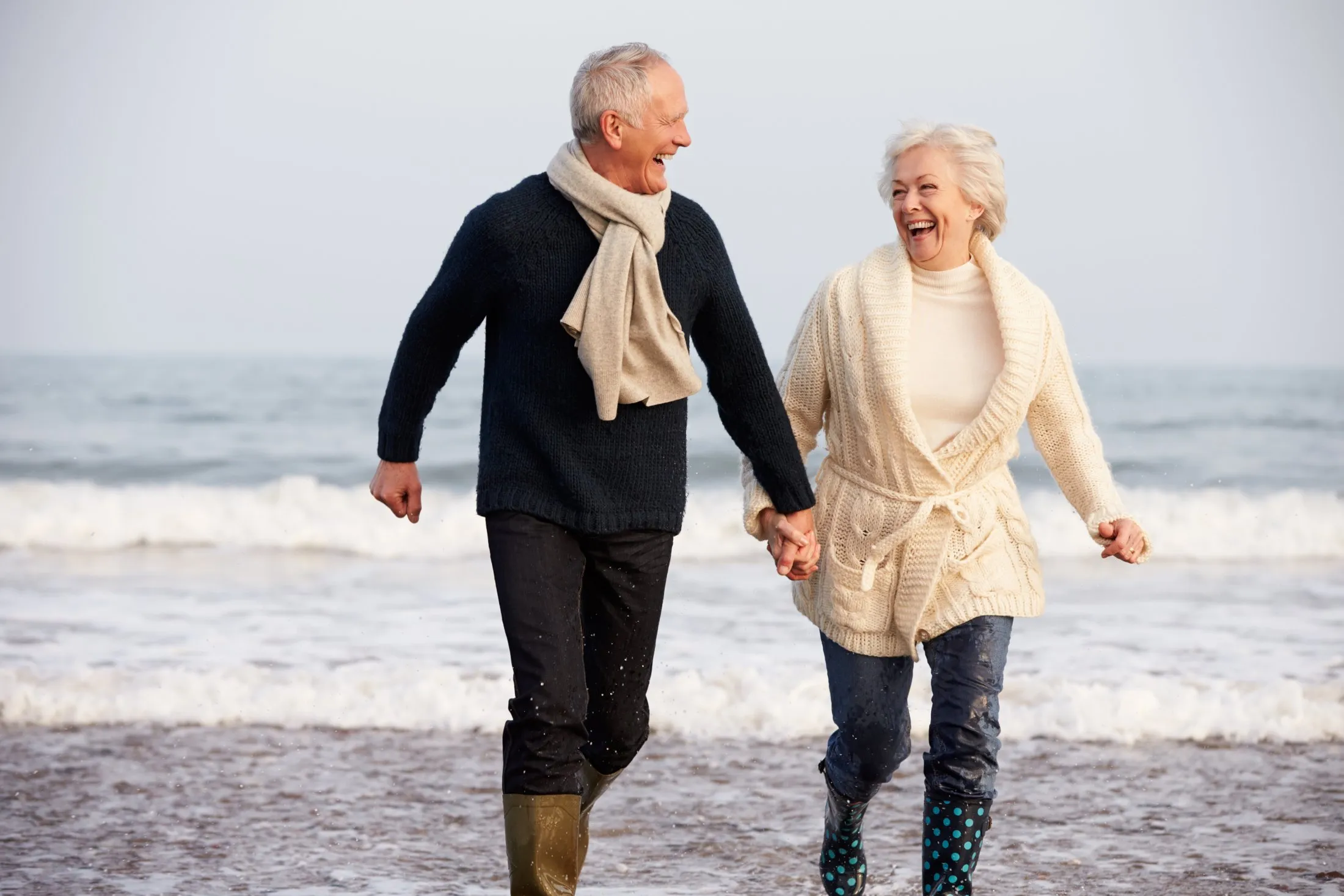 Happy elderly couple holding hands and walking on wet sand at the beach near ocean waves.