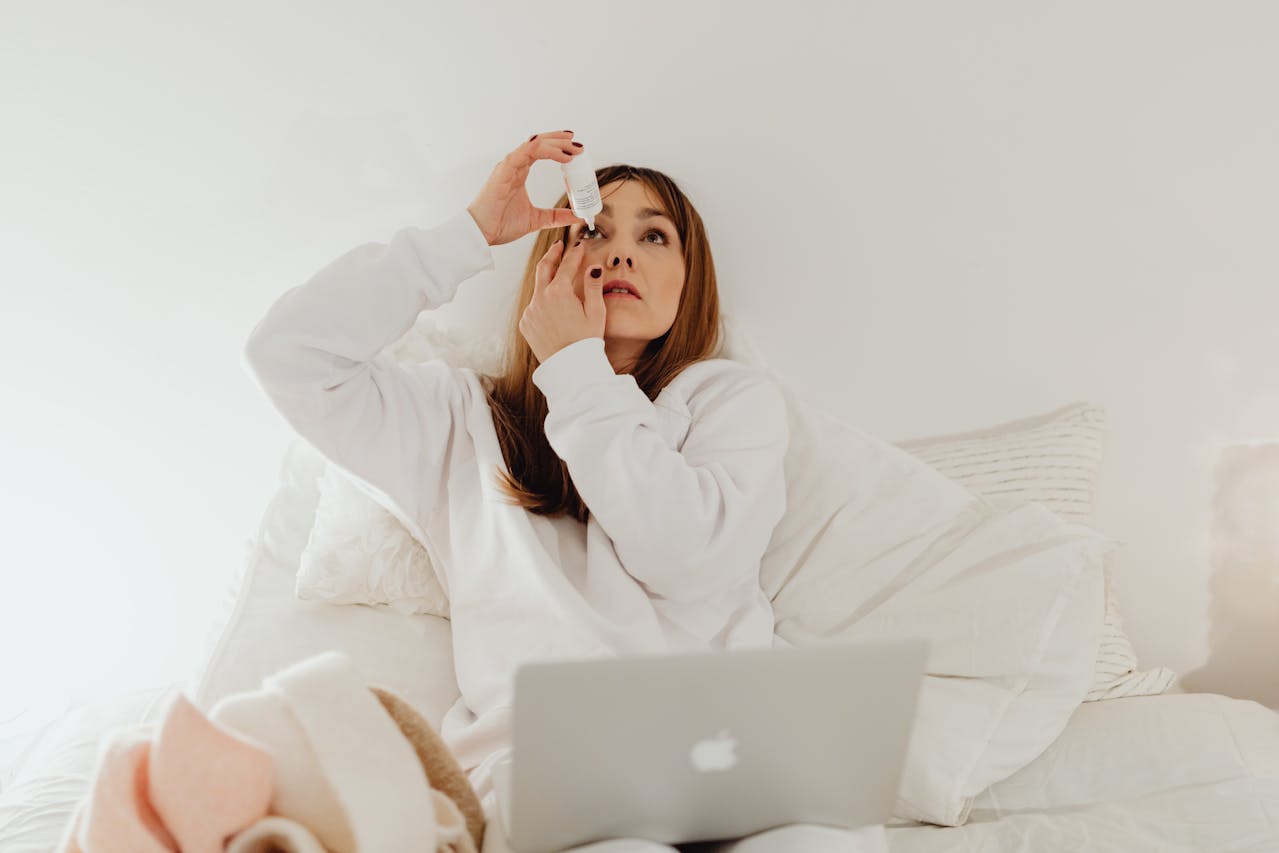 Woman sitting on bed, applying eye drops to her right eye with a laptop on her lap.