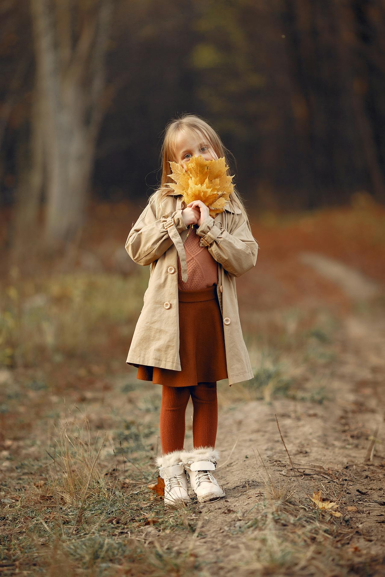 Young girl in beige coat holding yellow autumn leaves, standing on a dirt path in fall woods.