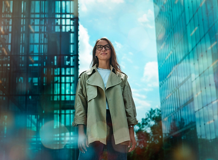 Confident young woman in glasses and green jacket standing outdoors with modern glass buildings behind her.