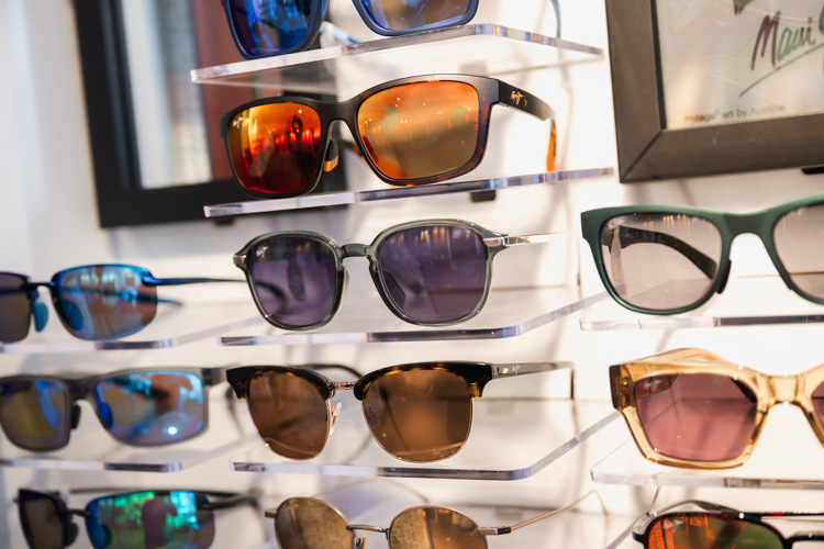 Variety of sunglasses with colored lenses displayed on clear shelves in an optical shop.