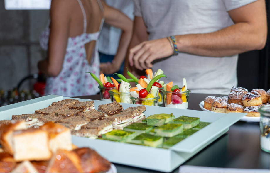 Angebot an Fingerfood mit Vollkorn-Sandwiches, Gemüsesticks in kleinen Gläsern, mit Paprika, Gurke und Kirschtomaten, und kleinen Gebäckstücken auf Tellern, Menschen im Hintergrund.