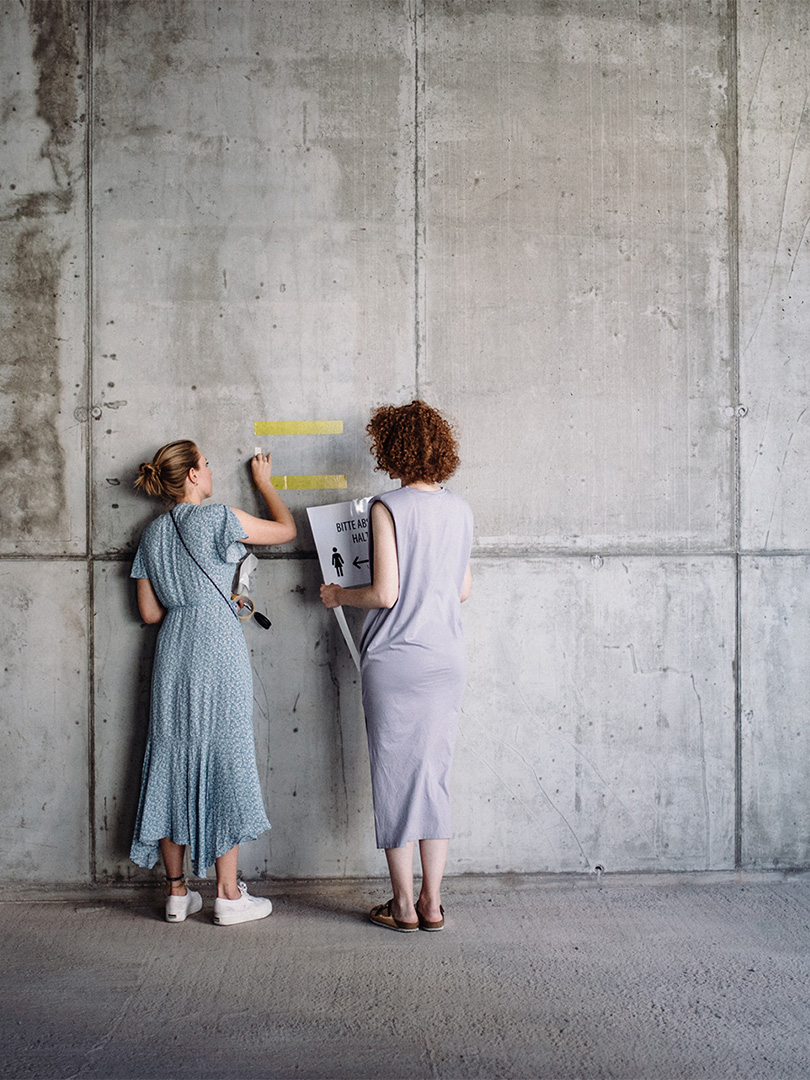 Zwei Frauen kleben gelbe Streifen an eine große graue Betonwand; eine Frau hält ein Schild mit der Aufschrift "Bitte Abstand halten" mit einem Richtungspfeil.