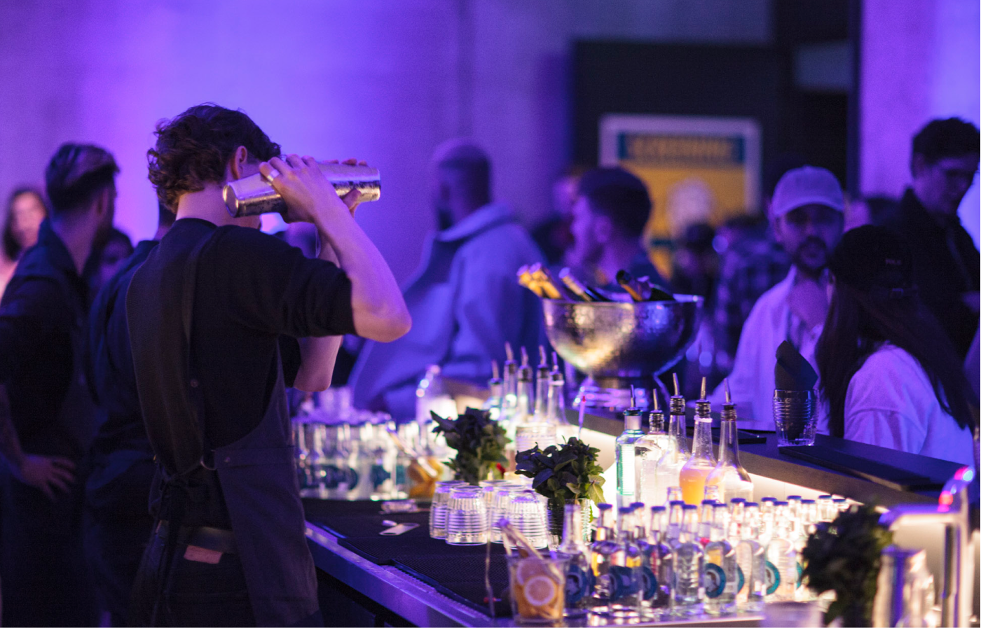Barkeeper mit schwarzem Hemd und Schürze schüttelt einen Cocktailshaker an einer Bar mit verschiedenen Flaschen, Gläsern und Eis, umgeben von Menschen in lila-blauem Licht.