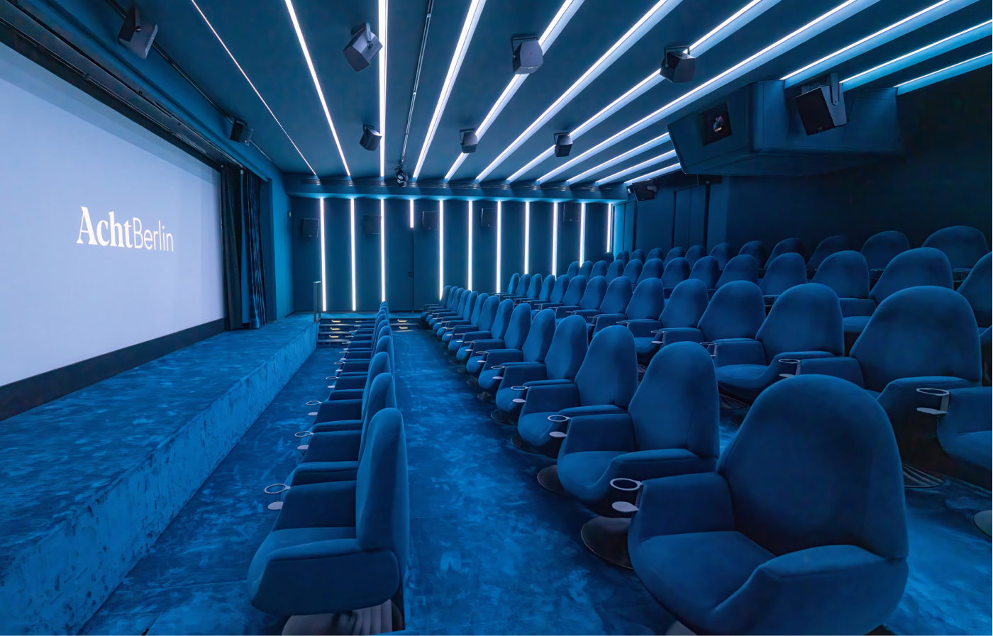 Kino mit modernem Design, blauen Samtsesseln mit Armlehnen und Getränkehalter, beleuchteter Decke mit linearen LED-Leuchten und großer Leinwand mit Schriftzug AchtBerlin.
