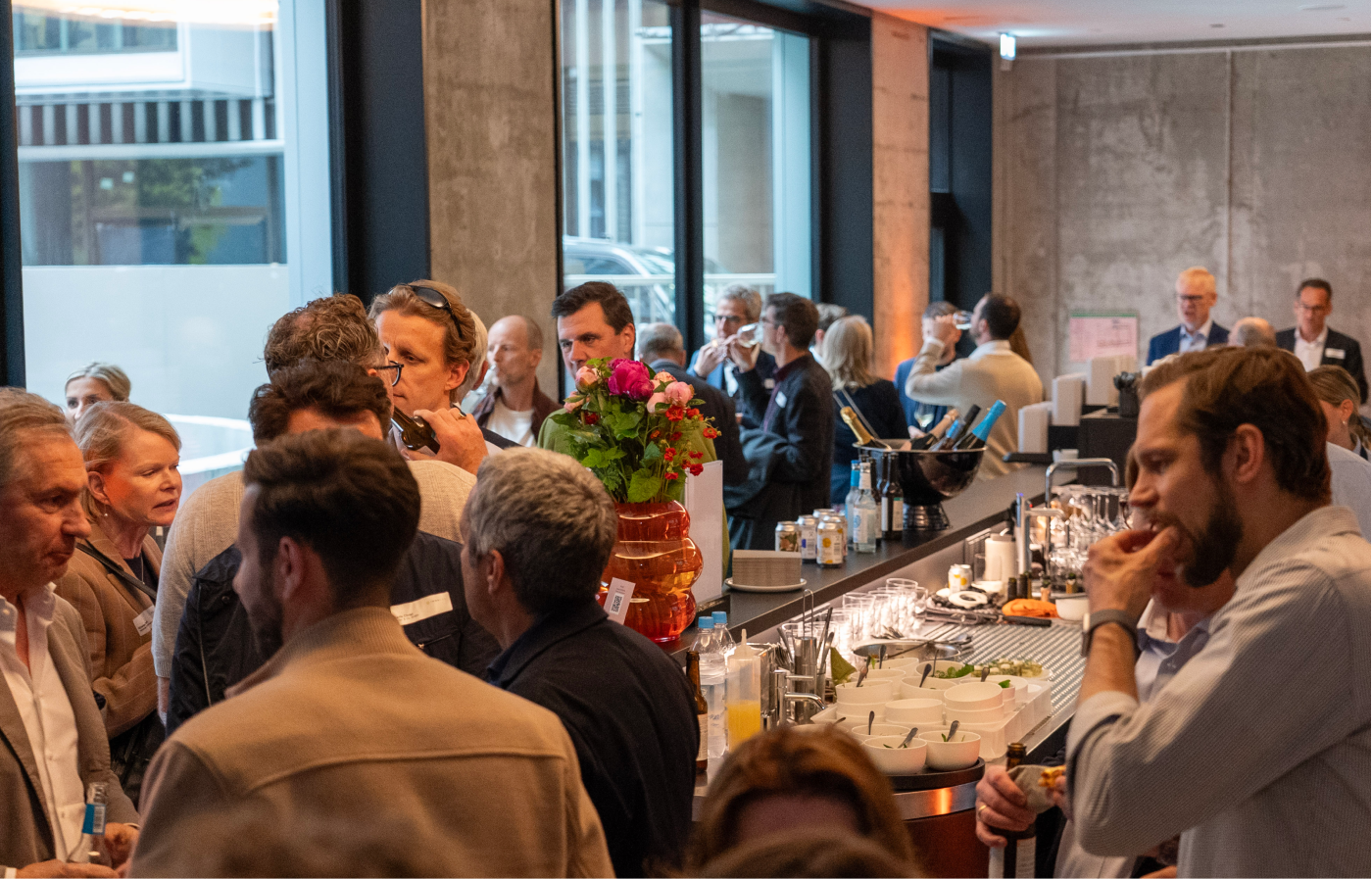 Menschen unterhalten sich und trinken bei einer Veranstaltung in einem modernen Innenraum mit großen Fenstern und Betonwänden, am langen Buffet mit Getränken, Gläsern und Snacks.