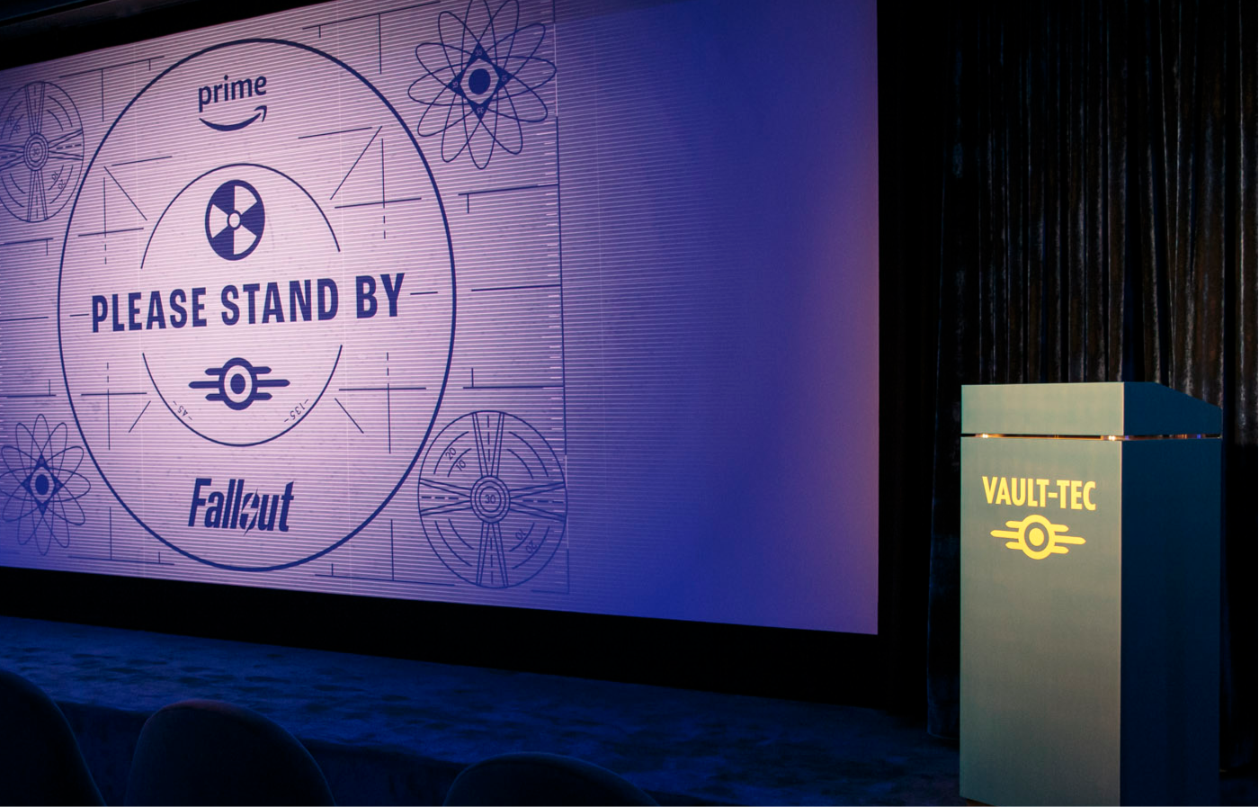 Ein dunkler Vorführraum mit einer großen Projektion der Fallout Spielserie, die eine Testbild-Anzeige mit der Aufschrift 'Please Stand By' zeigt; rechts steht ein beleuchtetes Podium mit dem Schriftzug 'Vault-Tec'.