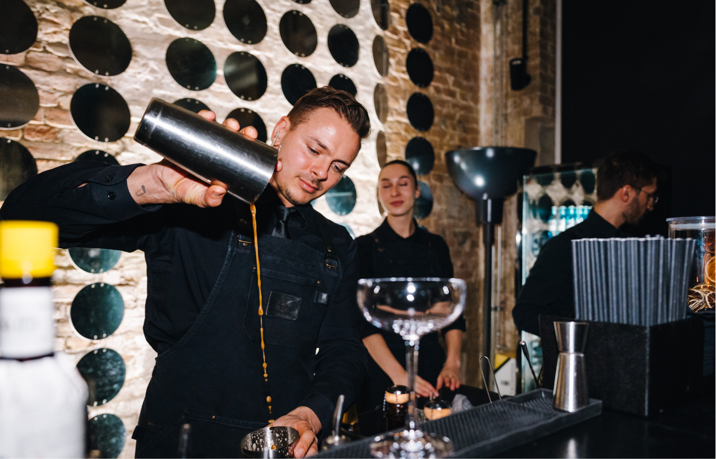 Barkeeper gießt mit einem Shaker einen Cocktail in ein Glas, während zwei weitere Mitarbeiter im Hintergrund an der Bar stehen, Umgebung mit runden Spiegeln an einer Backsteinwand.