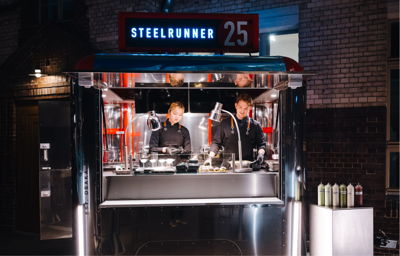 Zwei Köche in schwarzer Arbeitskleidung arbeiten in einem modernen, metallischen Foodtruck mit der Aufschrift STEELRUNNER 25 bei Nacht.