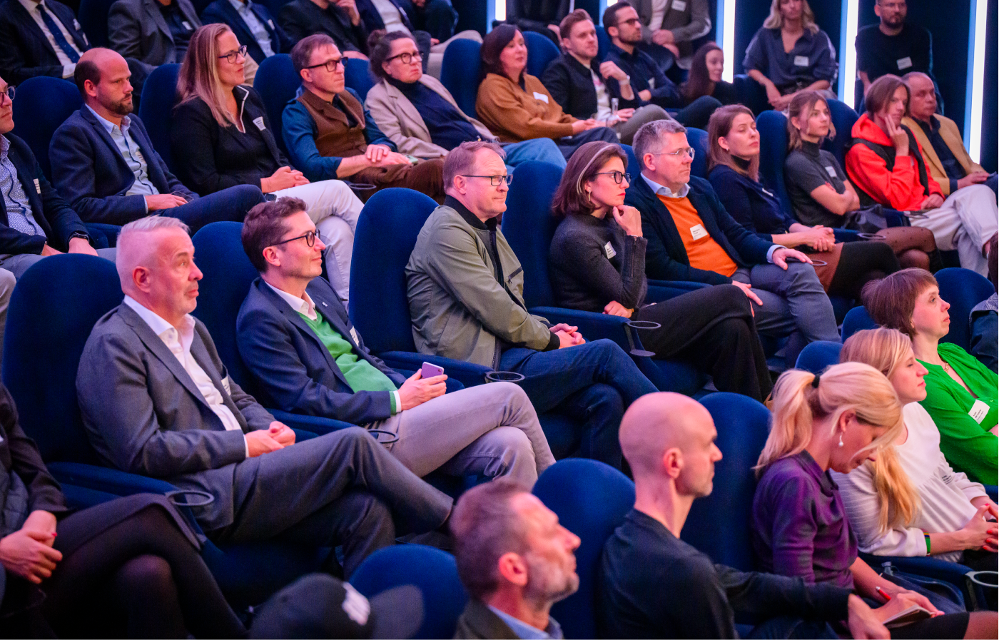Mehrere Personen sitzen in einem Auditorium auf blauen Sesseln und hören aufmerksam zu.