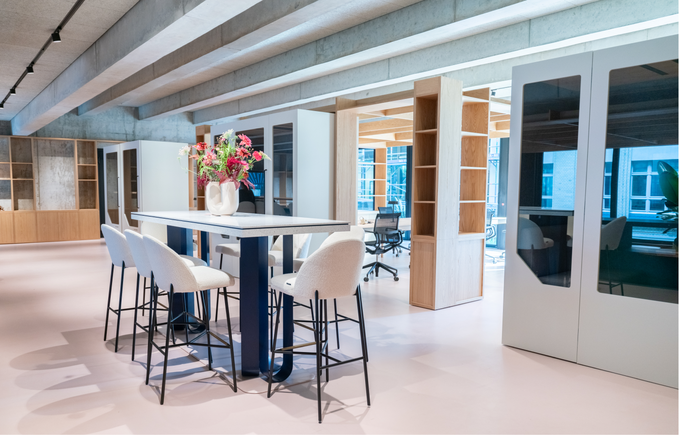 Moderner Coworking-Space mit langem Tisch und fünf gepolsterten Stühlen, darüber ein Blumenarrangement; im Hintergrund Holzmöbel, Glaskabinen und große Fenster mit Blick auf ein Gebäude.