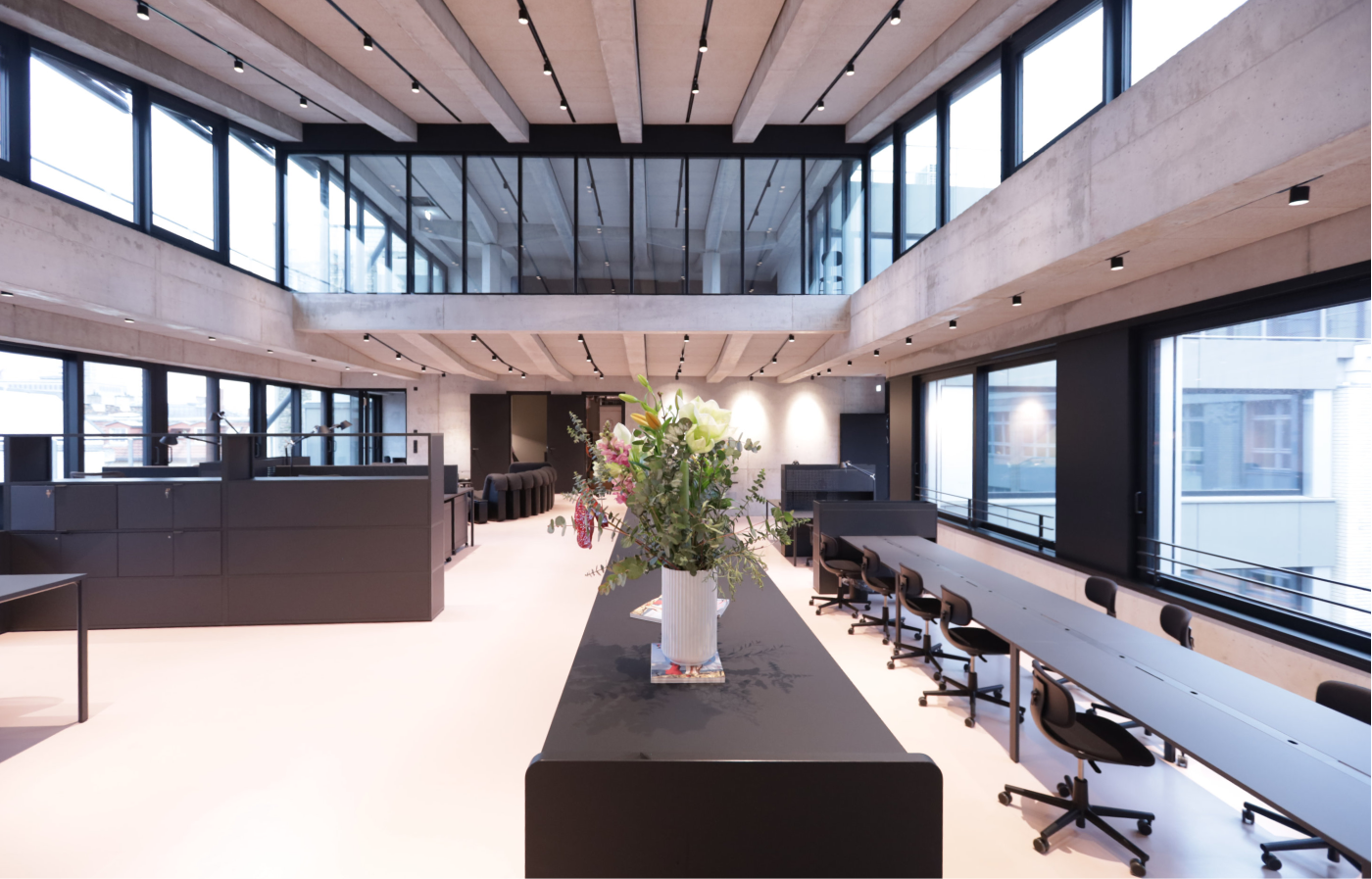 Moderne Bürofläche mit hohen Decken, großen Fenstern, schwarzen Schreibtischen und Stühlen, sowie einer langen Tischinsel mit Blumenarrangement in der Mitte.