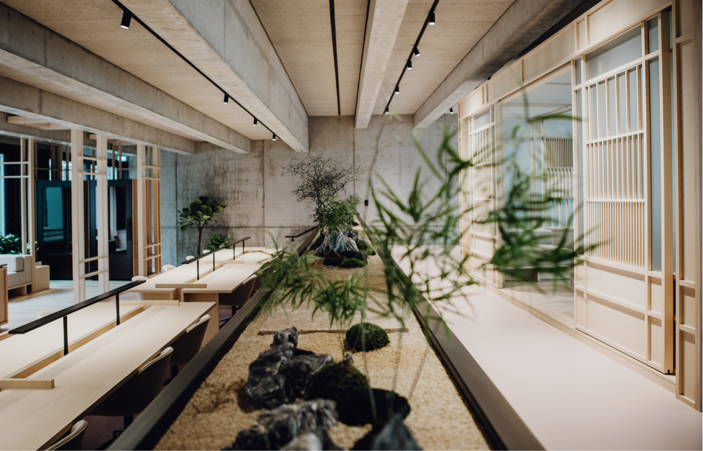 Moderner Innenraum mit schlichter Holz- und Betonoptik, langen Tischen mit Stühlen links und japanisch inspiriertem Zen-Garten mit Steinen und Pflanzen in der Mitte, rechts Wände mit Holzgitterelementen und Schiebetüren.