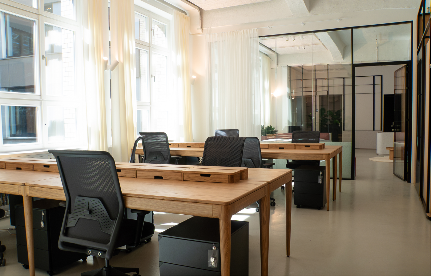 Helles, modernes Büro mit mehreren Holzschreibtischen, schwarzen ergonomischen Stühlen und viel natürlichem Licht durch große Fenster mit weißen Vorhängen, teilweise durch Glastrennwände unterteilt.