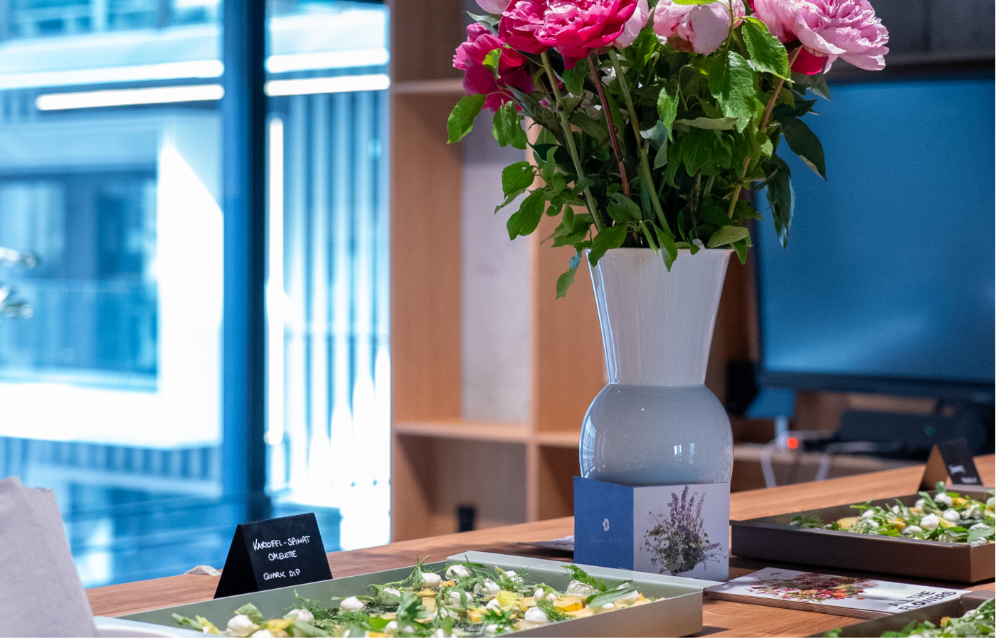 Ein weißer Keramikvasen mit pinken und rosa Blumen steht auf einem Holztisch neben Tabletts mit Salat und einer kleinen schwarzen Tafel mit der Beschriftung "Kartoffel-Spinat Omelette Quark Dip" in einem modernen Raum mit großen Fenstern.