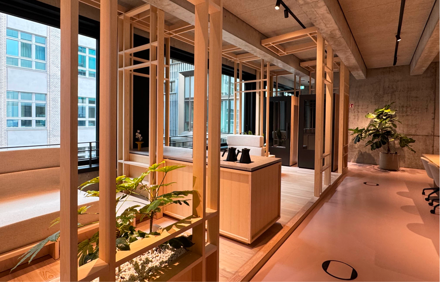 Modern office interior with wooden geometric partitions, beige seating area, plants in built-in planters and pots, large windows showing neighboring buildings, and exposed concrete ceiling with track lighting.