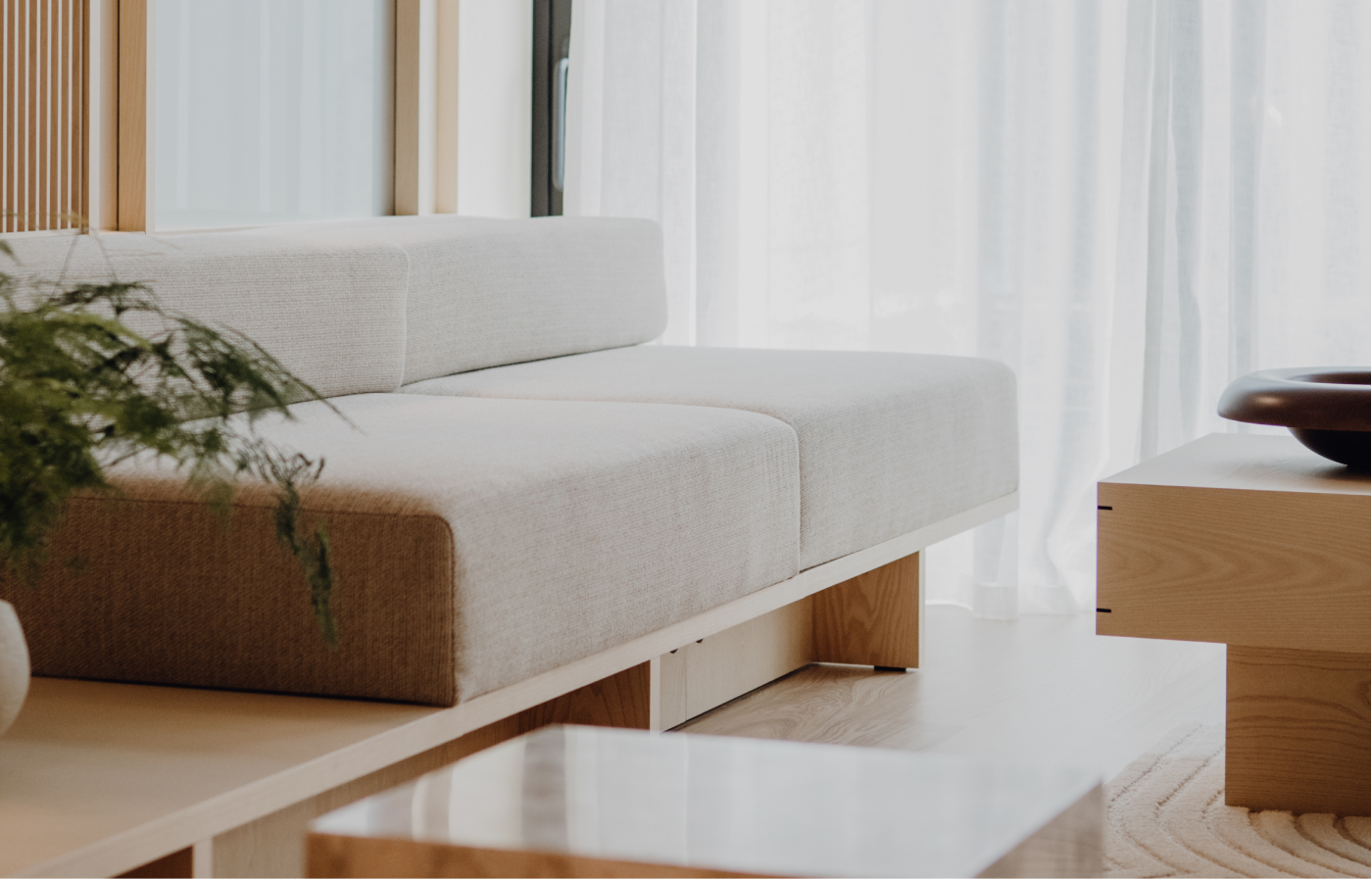 Helles modernes Wohnzimmer mit einem minimalistischen Sofa mit cremefarbenem Stoffbezug, einem Holztisch und einem Holzmöbel, dahinter weiße durchsichtige Vorhänge, ein grüner Topfpflanze links.