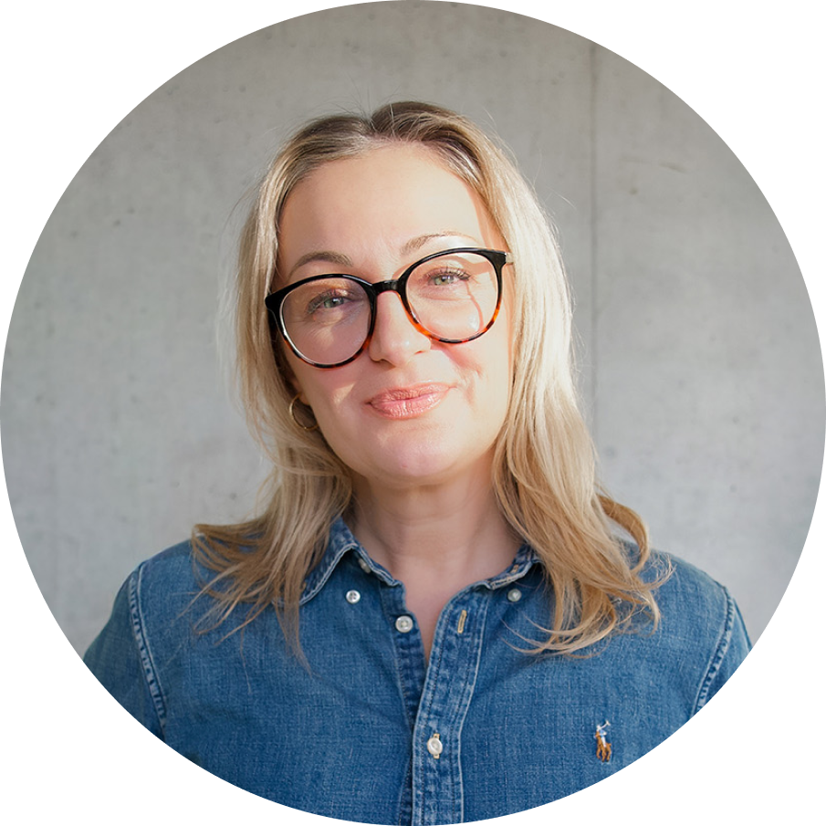 Frau mit langen blonden Haaren, schwarzer Brille und blauem Jeanshemd vor einer grauen Wand.