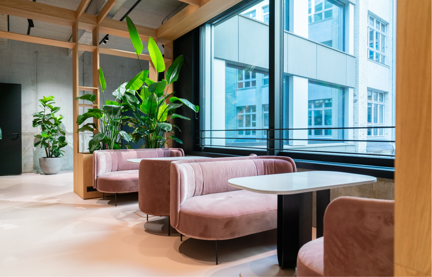 Modern coworking space with pink velvet bench seating and white tables by large windows, decorated with green plants and wooden beams.