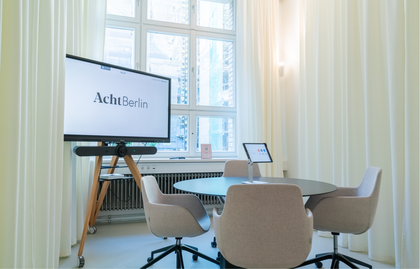 Moderner Besprechungsraum mit rundem schwarzen Tisch, vier grauen Bürostühlen auf Rollen, großem Fenster mit Blick auf ein Gebäude und Bildschirm auf einem Holz-Stativ mit der Aufschrift AchtBerlin.