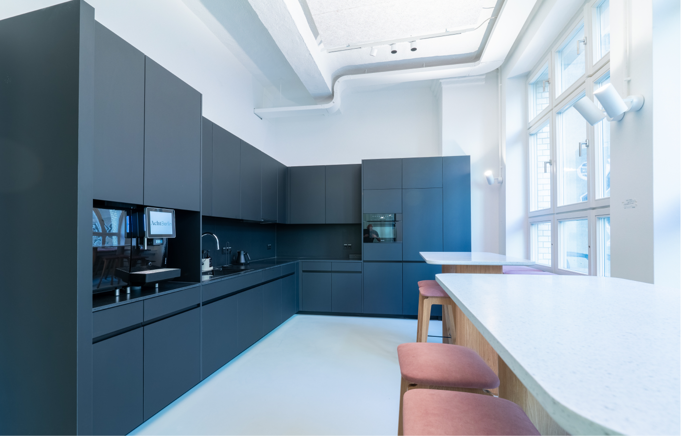 Moderne Küchenzeile mit mattschwarzen Schränken, integriertem Kaffeeautomaten und eingebautem Ofen in hell beleuchtetem Raum mit großen Fenstern und Stehtischen mit rosa Sitzhockern.