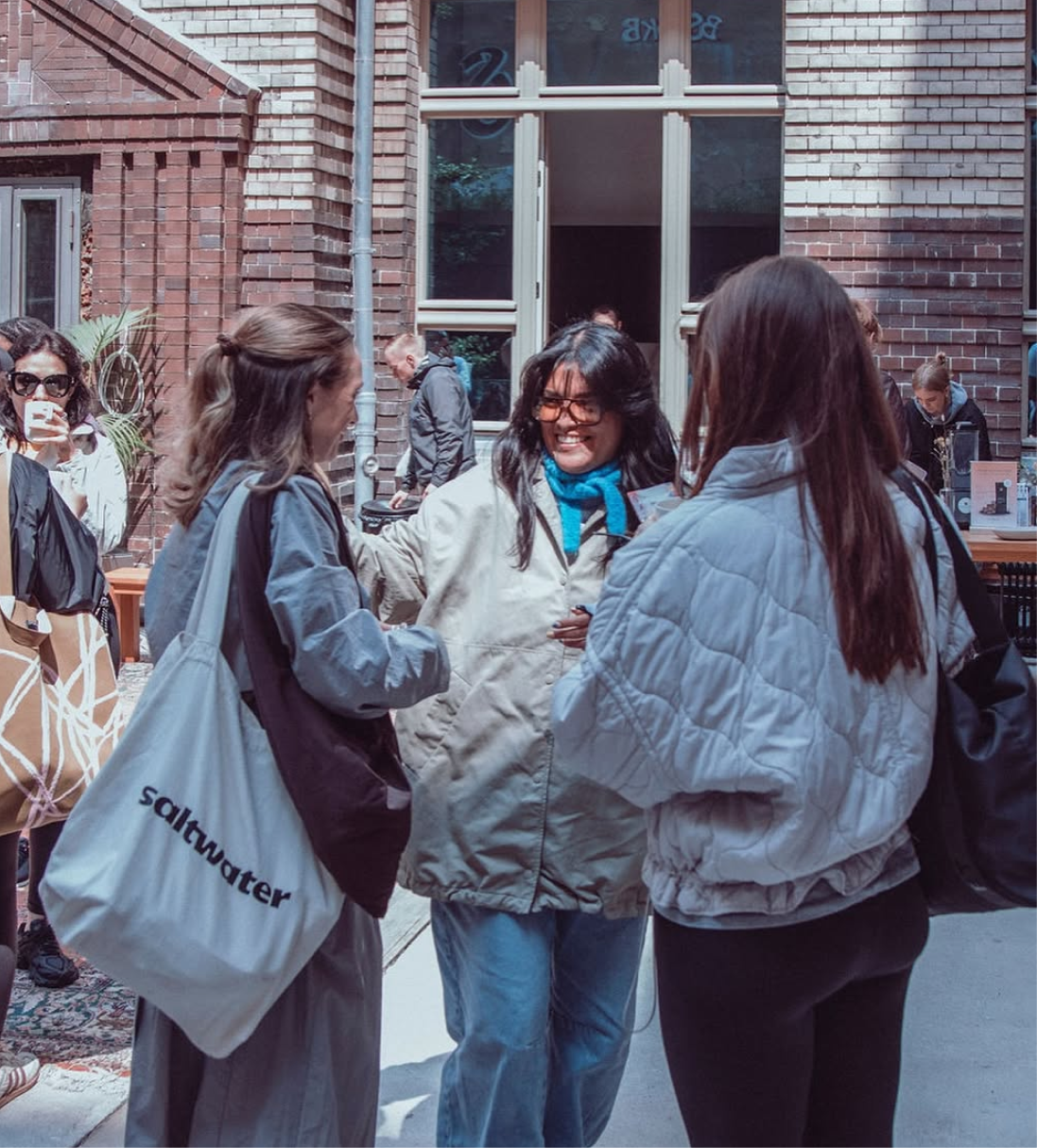 Drei Frauen unterhalten sich fröhlich im Freien vor einem Backsteingebäude, eine trägt eine Tasche mit der Aufschrift »saltwater«.