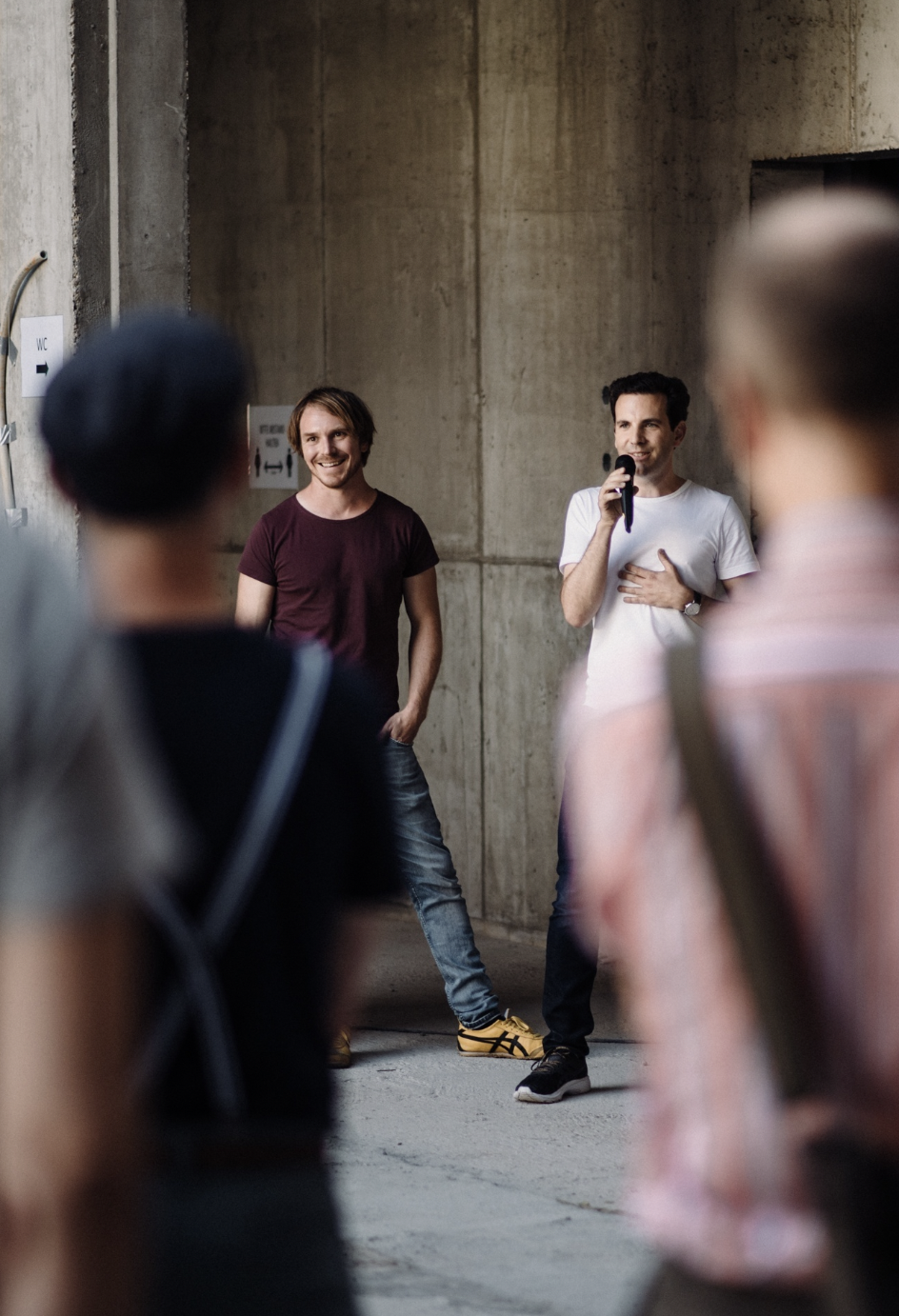 Zwei Männer stehen vor einer Betonwand, einer in weißem T-Shirt spricht in ein Mikrofon, der andere in dunkelrotem T-Shirt lächelt, während zwei unscharfe Personen im Vordergrund stehen.