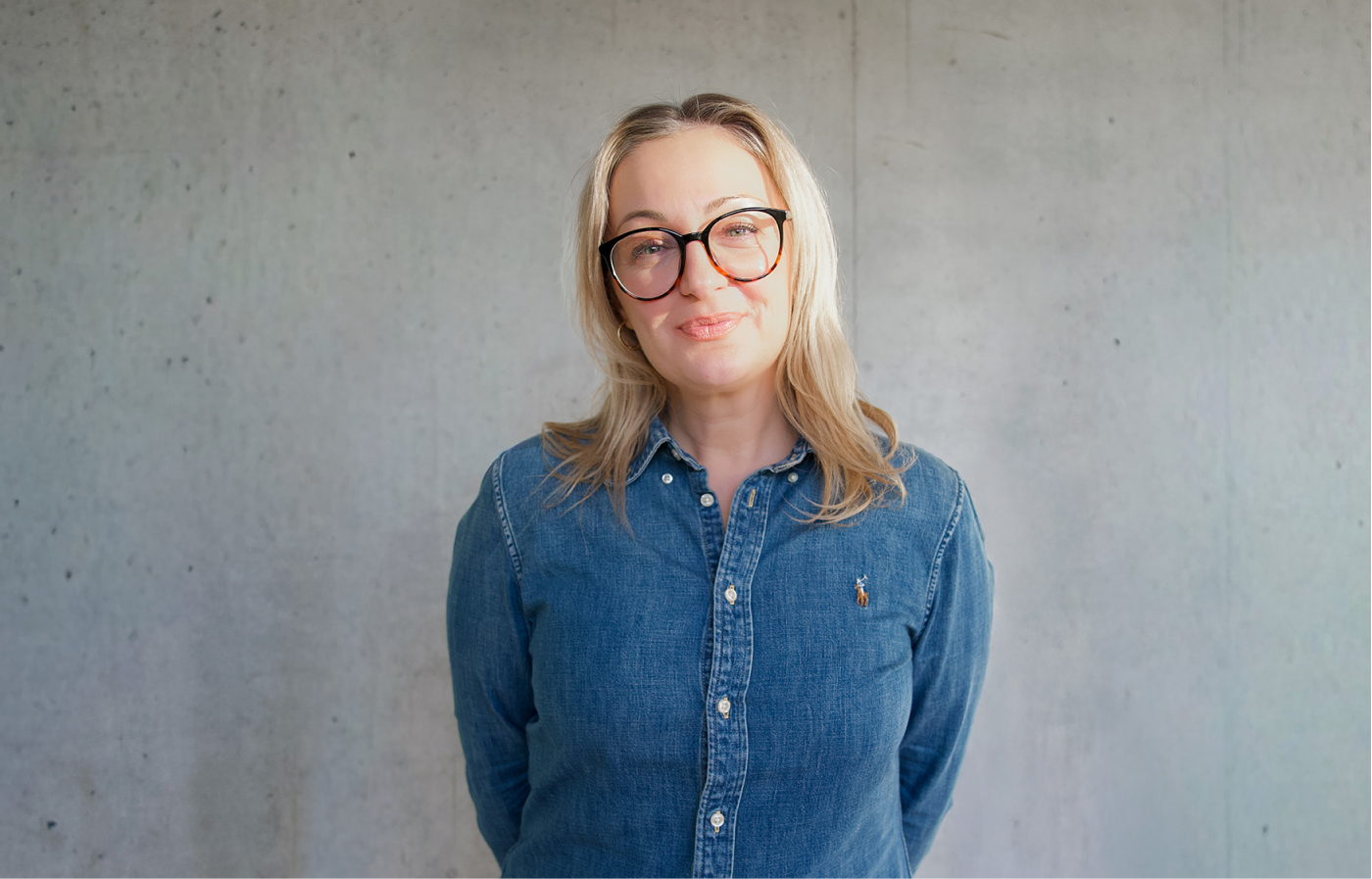 Frau mit blonden Haaren, die schwarze Brillengestelle trägt und ein blaues Jeanshemd vor einer grauen Betonwand steht.