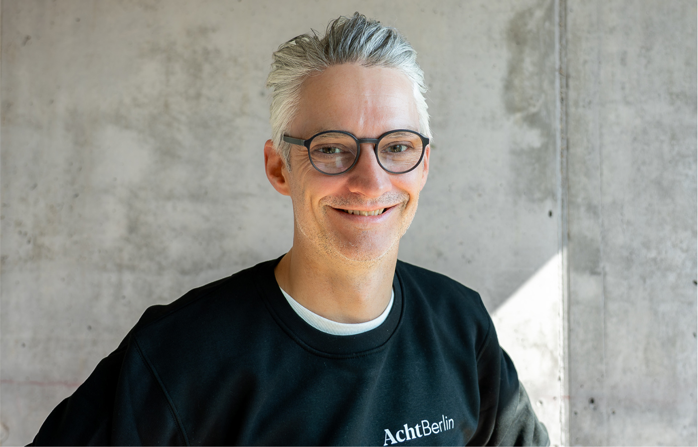 Mann mit grauen Haaren und Brille trägt schwarzen Pullover mit der Aufschrift AchtBerlin vor einer hellgrauen Betonwand.