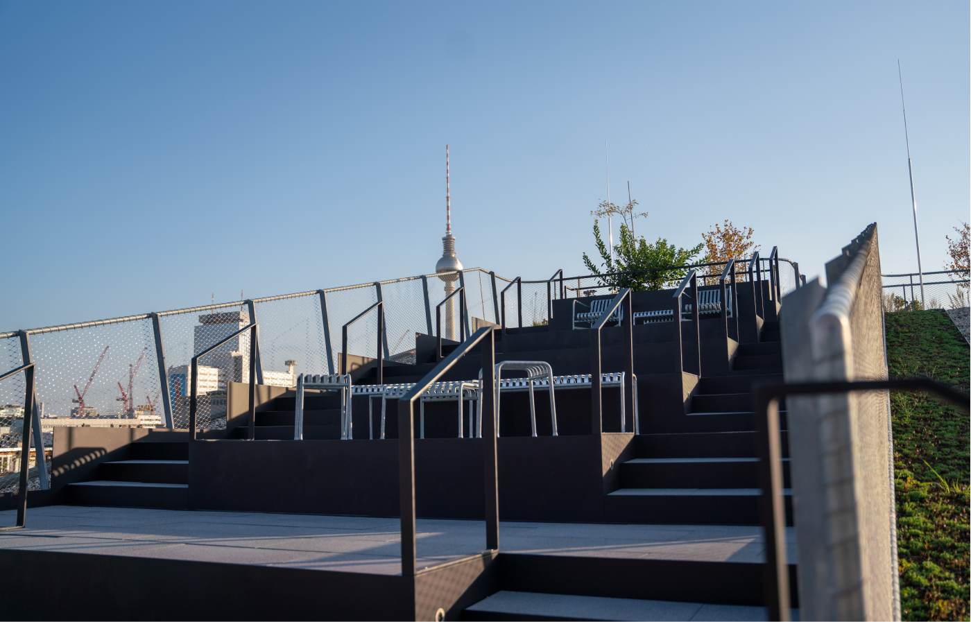 Moderne Außentreppe mit schwarzen Stufen, Metallgeländer und Sitzgelegenheiten aus Metall in einer offenen Umgebung mit klarem blauen Himmel und Blick auf den Fernsehturm in Berlin im Hintergrund.