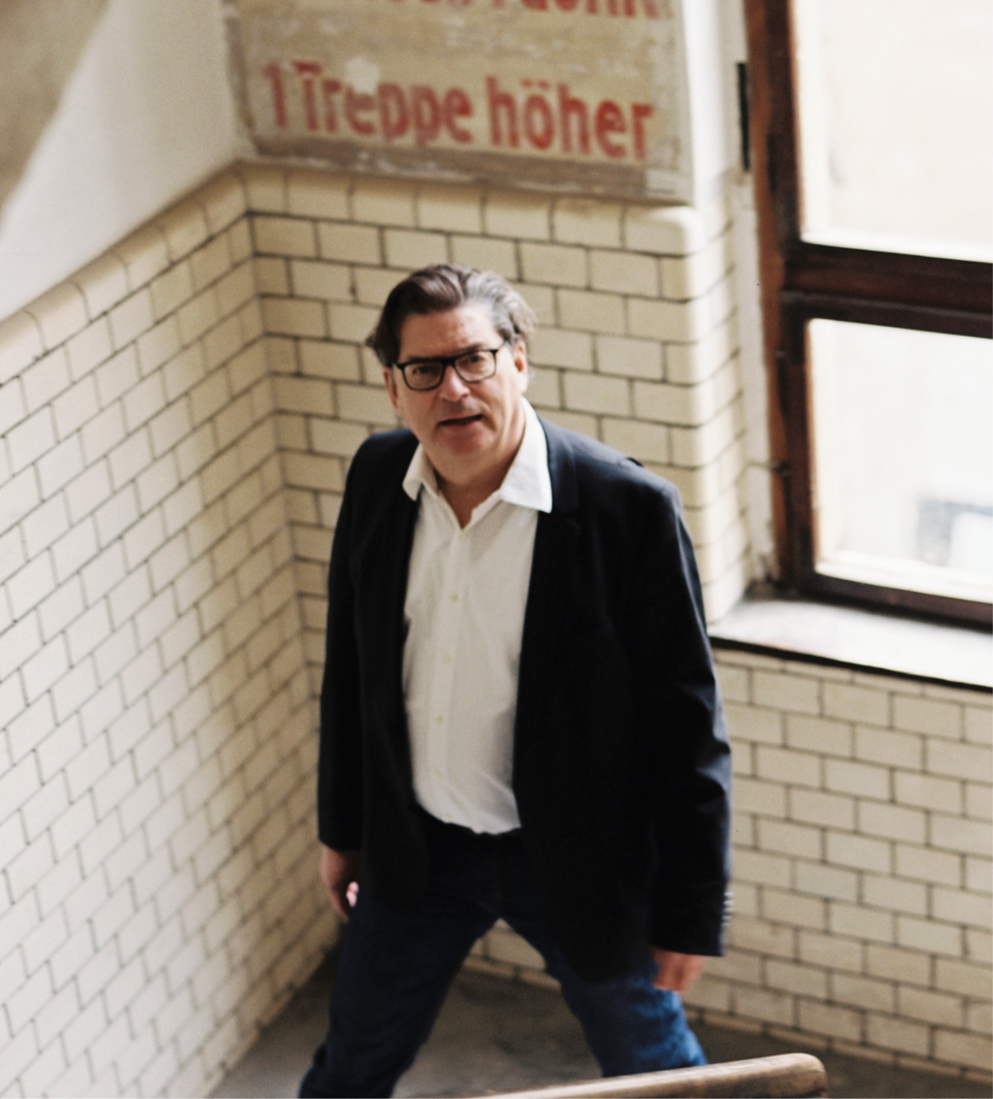 Mann mit Brille, weißem Hemd und schwarzem Sakko geht eine Treppe mit weißen Kacheln hinauf neben einem Fenster, im Hintergrund ein Schild mit Schriftzug ‚1 Treppe höher‘.