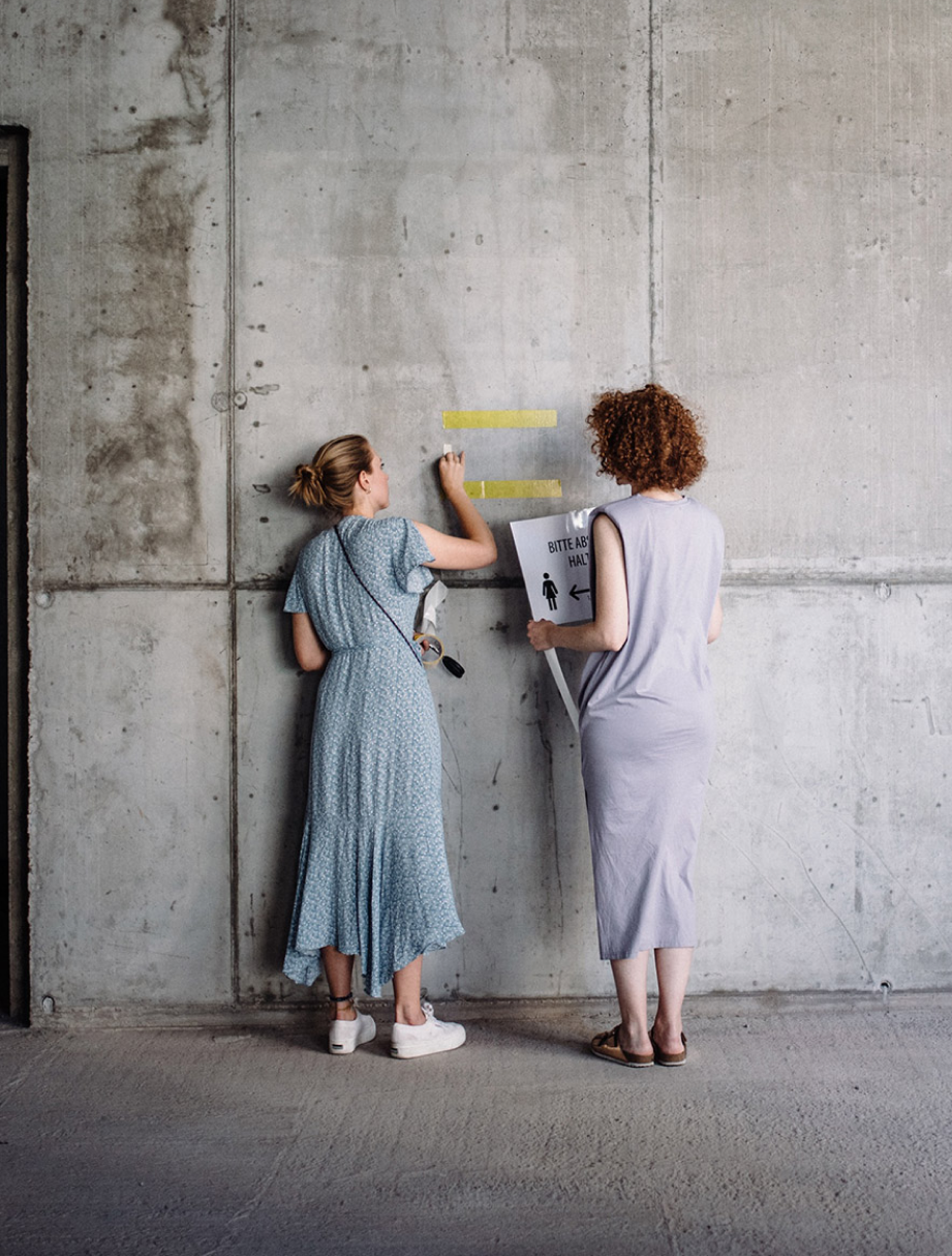 Zwei Frauen kleben gelbe Markierungen an eine Betonwand; eine hält ein Schild mit der Aufschrift ‚Bitte Abstand halten‘ und einem Piktogramm einer Person mit Pfeil nach links.
