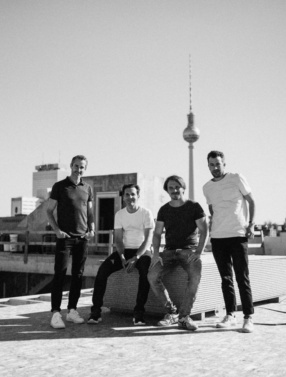 Schwarzweiß-Foto von vier jungen Männern auf einem Dach mit dem Berliner Fernsehturm im Hintergrund, zwei stehen und zwei sitzen auf einer Bank.