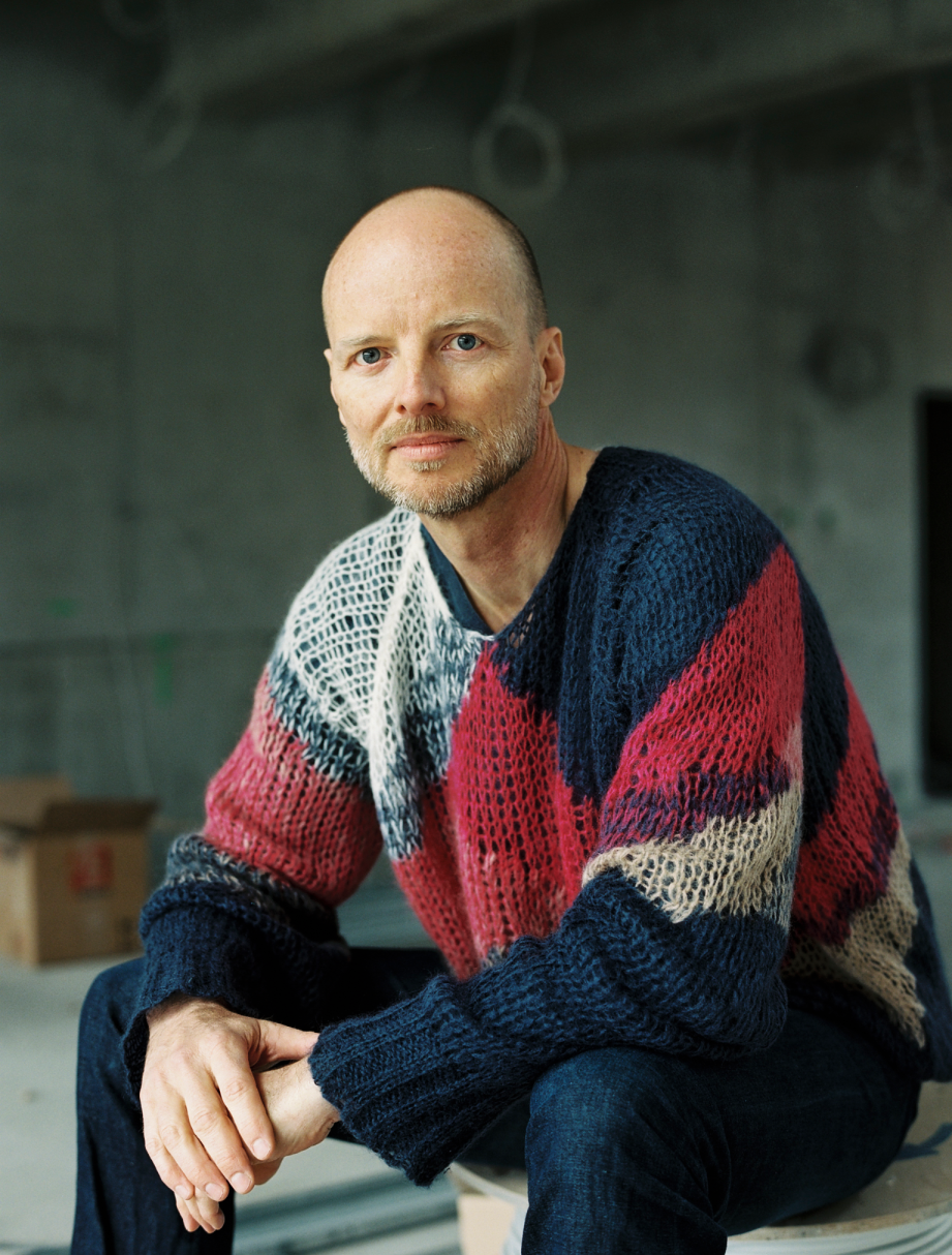 Porträt eines sitzenden Mannes mit Glatze und kurzem Bart, der einen mehrfarbigen, grobmaschigen Pullover in Rot, Blau und Beige trägt, vor einem unscharfen, grauen Hintergrund.