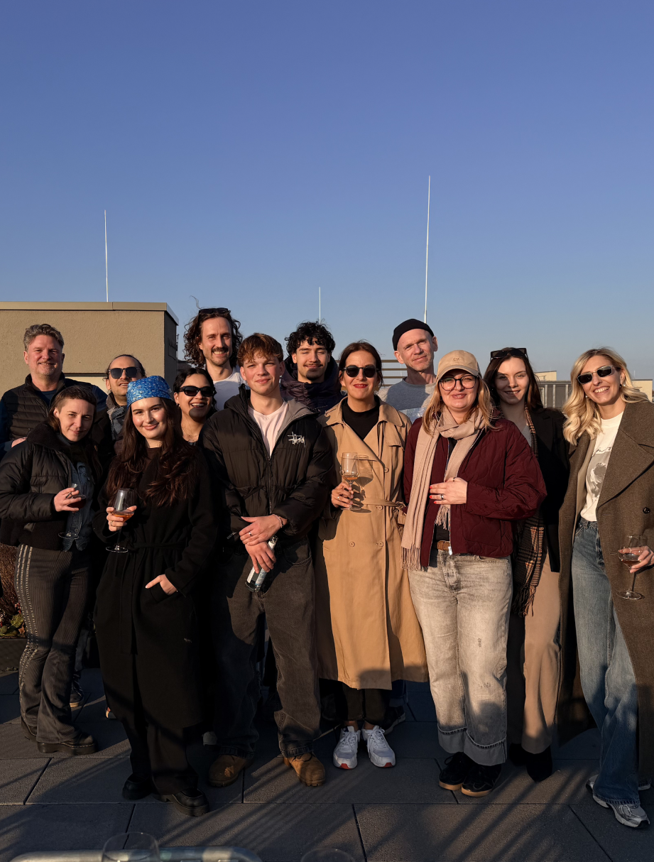 Eine Gruppe von zwölf Menschen steht eng zusammen auf einer Dachterrasse bei sonnigem Wetter und blauem Himmel. Die meisten tragen Herbst- oder Winterkleidung wie Jacken, Mäntel und Schals. Einige halten Weingläser oder eine Flasche in den Händen. Alle blicken lächelnd zur Kamera.