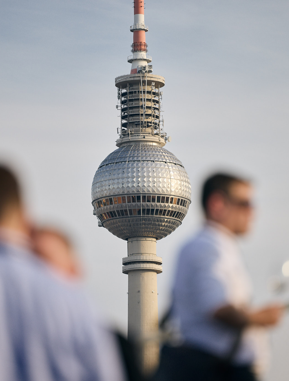 Nahaufnahme des Fernsehturms Berlin mit unscharfen Menschen im Vordergrund, die eine geschäftliche Atmosphäre vermitteln.