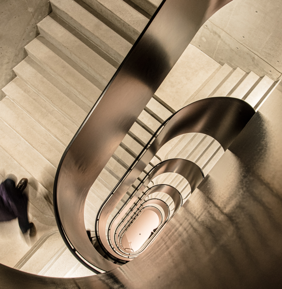 Blick nach unten auf eine moderne, helle Wendeltreppe mit glattem schwarzen Geländer und hellen Stufen aus Beton. Eine Person mit dunkler Kleidung steigt gerade die Treppe hinab.