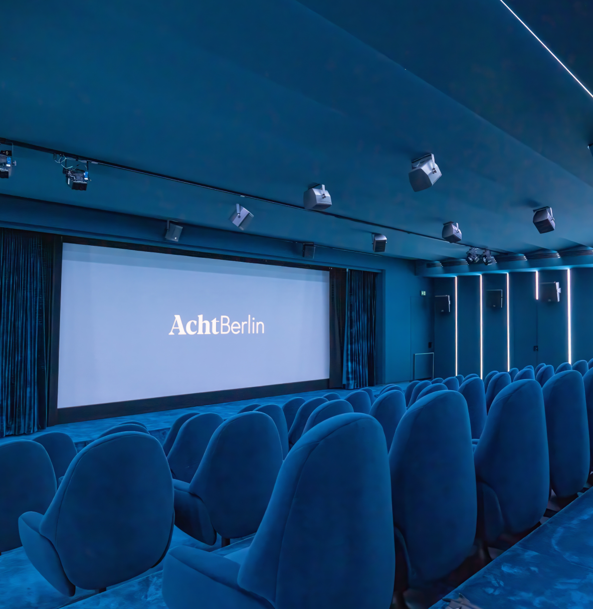 Modernes Kino mit samtblauen Sitzen und einer großen Leinwand, auf der das Logo „AchtBerlin“ leuchtet.